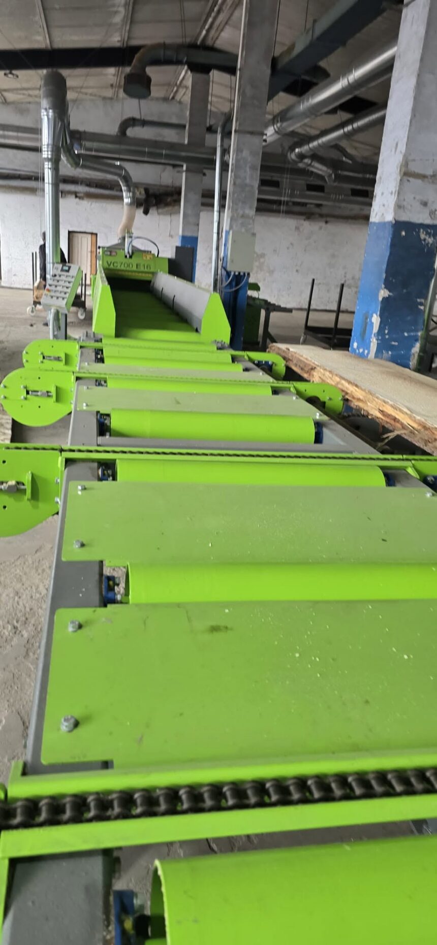 Green industrial conveyor system with rollers and guides in a dusty workshop, with overhead ducts and machinery in the background.