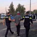 Two police officers in tactical gear escort a blurred detainee along a gravel street; a toppled white car is visible in the background.