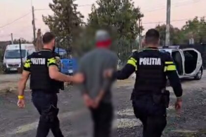 Two police officers in tactical gear escort a blurred detainee along a gravel street; a toppled white car is visible in the background.