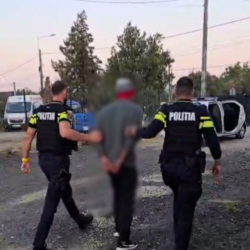 Two police officers in tactical gear escort a blurred detainee along a gravel street; a toppled white car is visible in the background.