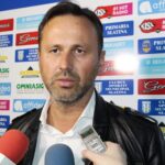 Man in a white shirt and black leather jacket being interviewed, with a sponsor-branded backdrop behind him and microphones nearby.