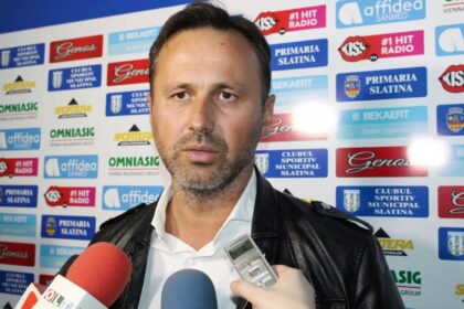 Man in a white shirt and black leather jacket being interviewed, with a sponsor-branded backdrop behind him and microphones nearby.
