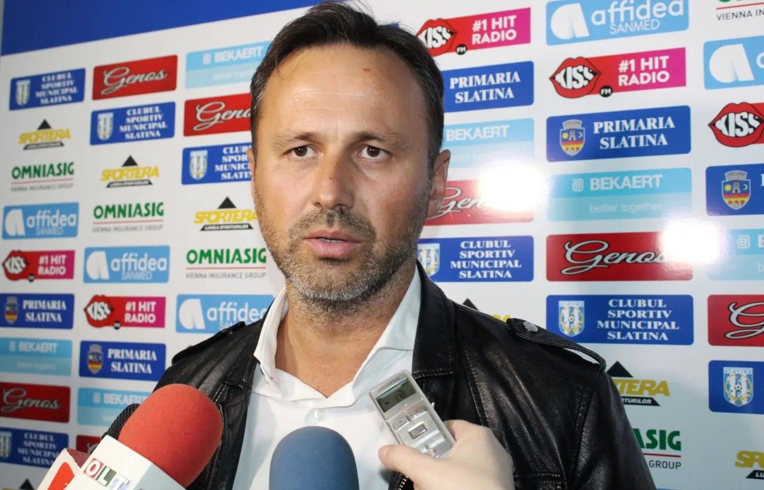 Man in a white shirt and black leather jacket being interviewed, with a sponsor-branded backdrop behind him and microphones nearby.