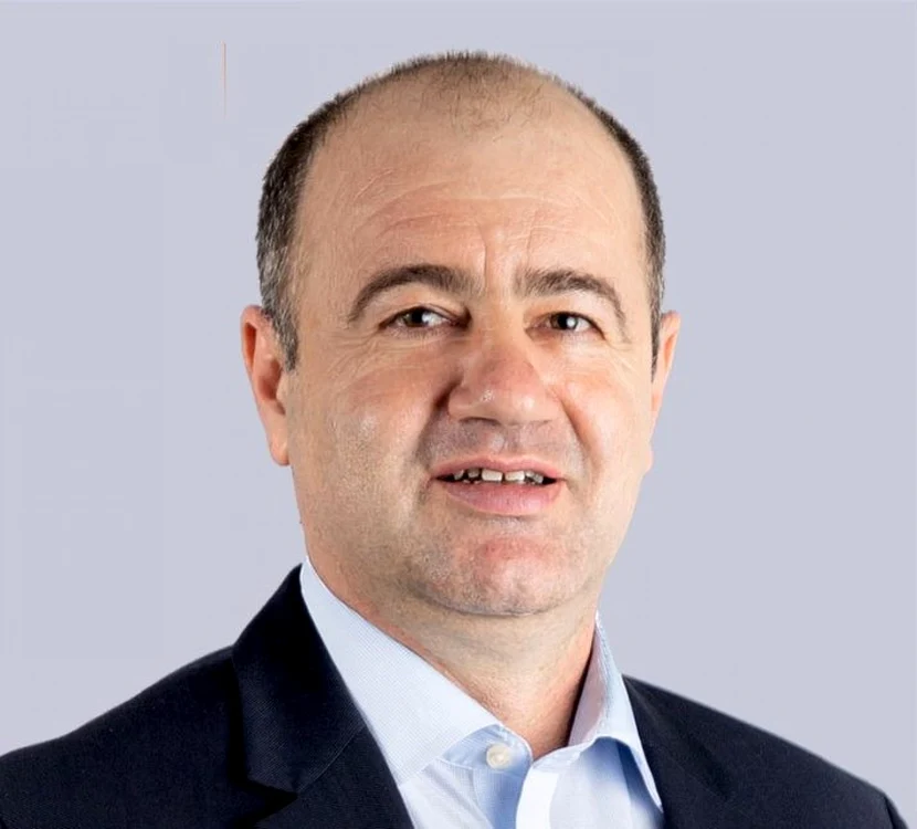 Head-and-shoulders portrait of a middle-aged man in a navy suit and light blue shirt against a gray background.
