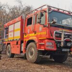 Red fire engine labeled 'POMPIERII' and '112' parked on rocky dirt with a firefighter nearby, autumn trees in the background.