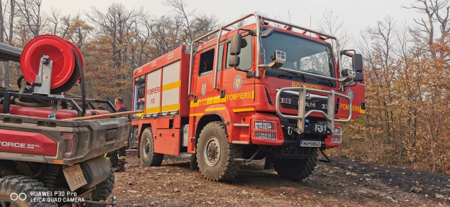 Red fire engine labeled 'POMPIERII' and '112' parked on rocky dirt with a firefighter nearby, autumn trees in the background.