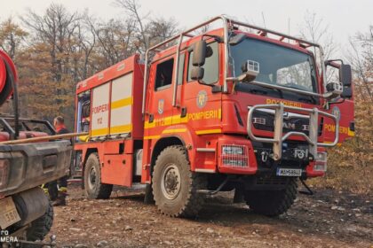 Red fire engine labeled 'POMPIERII' and '112' parked on rocky dirt with a firefighter nearby, autumn trees in the background.