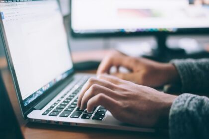 Person typing on a laptop at a desk with a second monitor in the background.