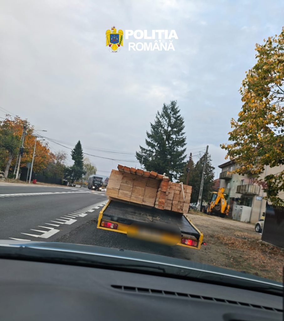 Yellow flatbed truck carrying a large load of stacked lumber on a street, with autumn trees and a construction excavator in the background, and a POLITIA ROMÂNĂ watermark overhead.