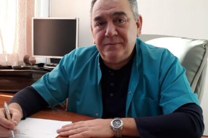 Man in a teal shirt signs a document at a wooden desk, with a computer monitor in the background.