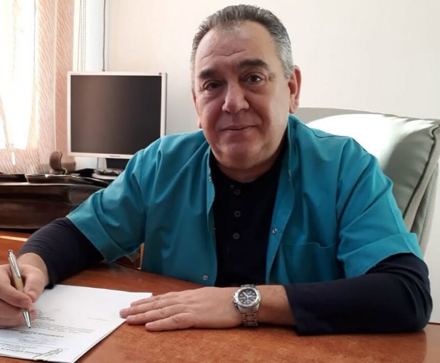 Man in a teal shirt signs a document at a wooden desk, with a computer monitor in the background.