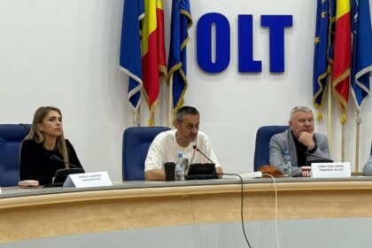Panel of three people at a conference table with microphones, in a room with flags and large blue 'OLT' letters behind them.