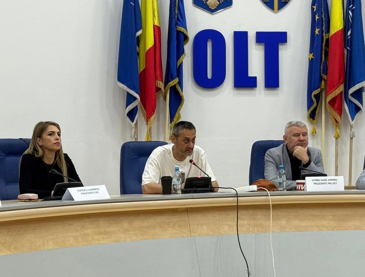Panel of three people at a conference table with microphones, in a room with flags and large blue 'OLT' letters behind them.