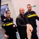 Three men in black jackets with yellow stripes stand indoors in a blurred group photo.
