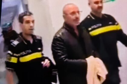 Three men in black jackets with yellow stripes stand indoors in a blurred group photo.