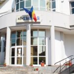 White medical clinic building with glass doors, columns, Romanian flag, and Red Cross sign; steps with potted plants under a blue sky