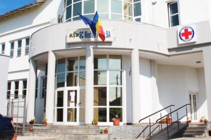 White medical clinic building with glass doors, columns, Romanian flag, and Red Cross sign; steps with potted plants under a blue sky