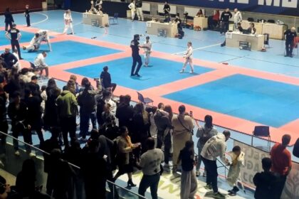 Indoor martial arts competition on blue and red mats; two young athletes sparring while an official stands nearby, with spectators along the edge and judges at tables in the background.