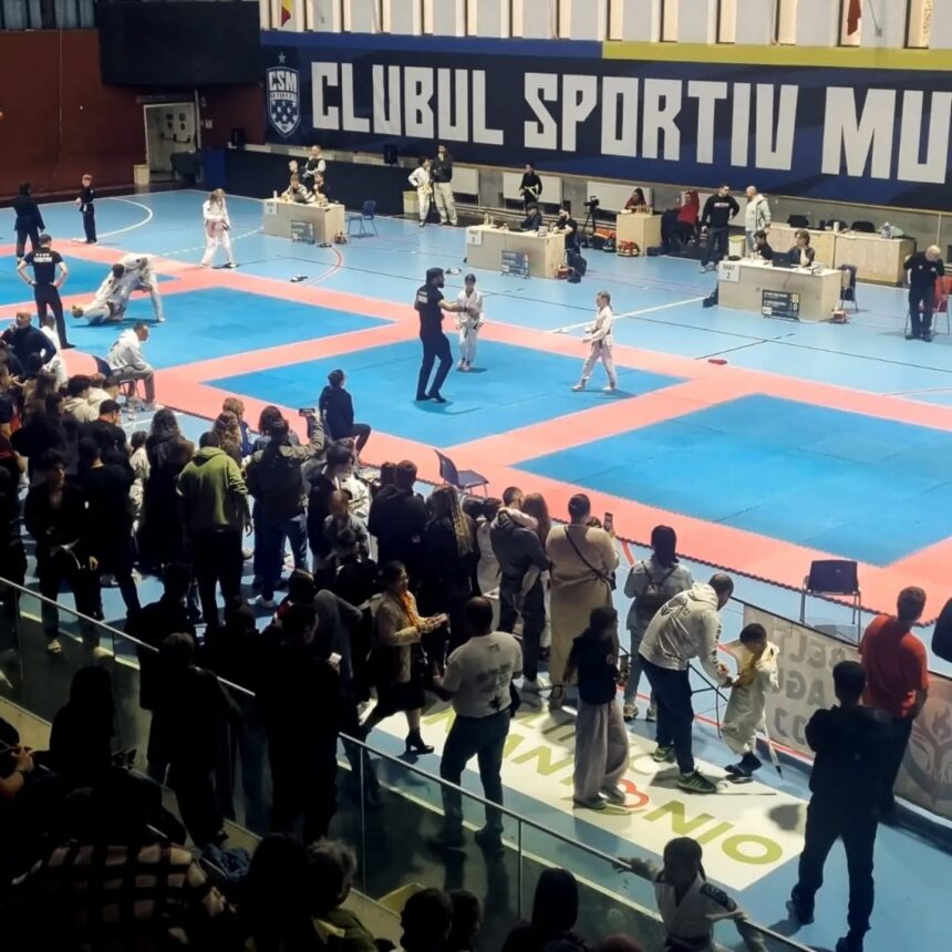 Indoor martial arts competition on blue and red mats; two young athletes sparring while an official stands nearby, with spectators along the edge and judges at tables in the background.