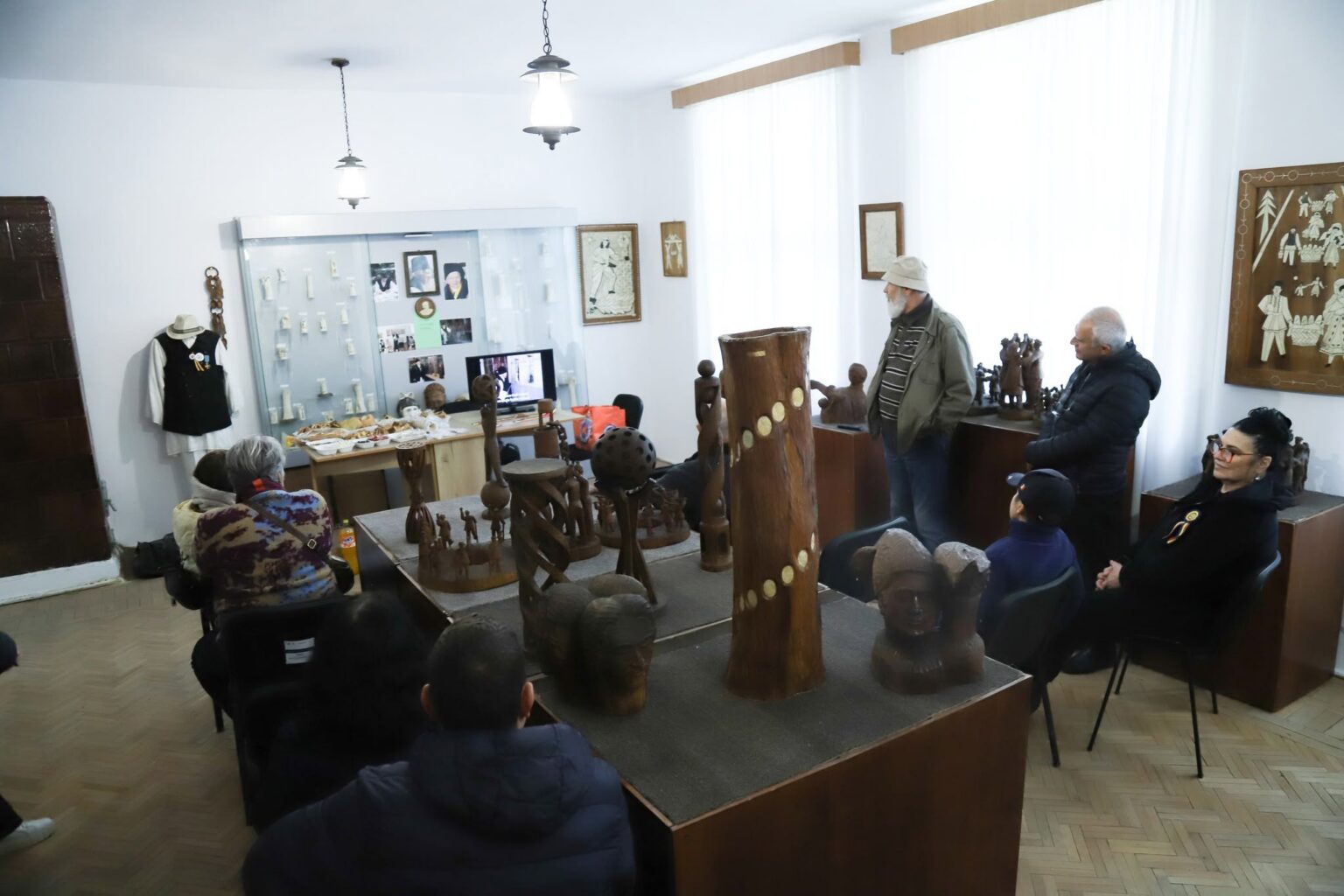 Group of people seated and standing in a museum room, viewing wooden sculptures and a display cabinet with photos and artifacts.