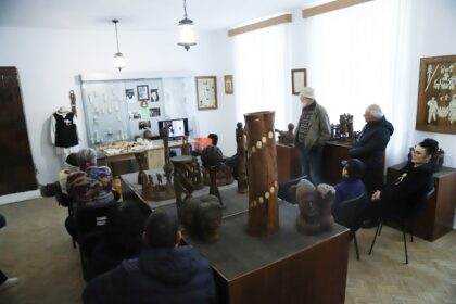 Group of people seated and standing in a museum room, viewing wooden sculptures and a display cabinet with photos and artifacts.