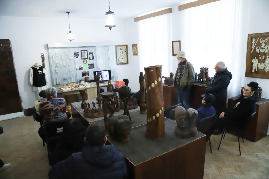 Group of people seated and standing in a museum room, viewing wooden sculptures and a display cabinet with photos and artifacts.