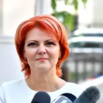 Woman with bright red hair speaking to reporters outdoors, microphones in foreground, neutral expression on her face.