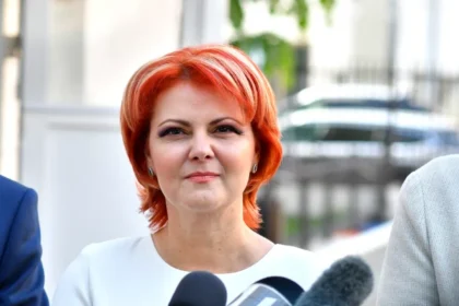 Woman with bright red hair speaking to reporters outdoors, microphones in foreground, neutral expression on her face.
