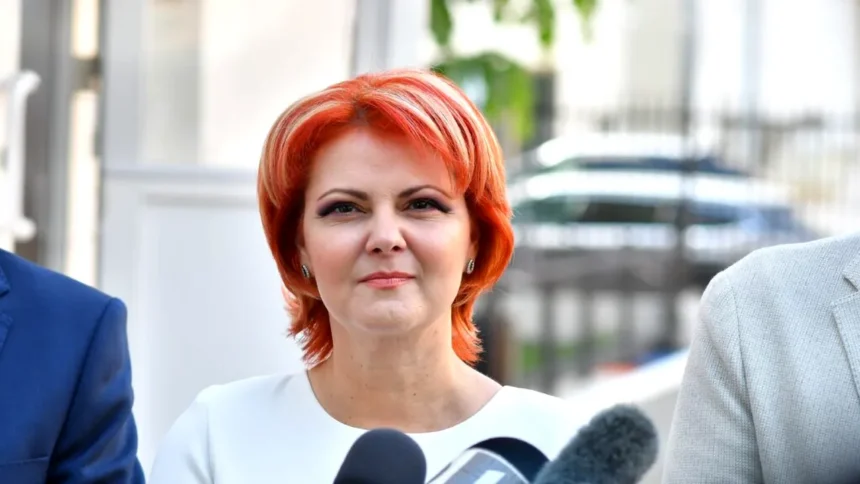 Woman with bright red hair speaking to reporters outdoors, microphones in foreground, neutral expression on her face.