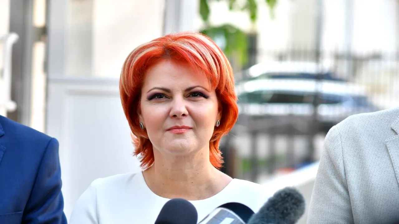 Woman with bright red hair speaking to reporters outdoors, microphones in foreground, neutral expression on her face.