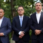 Three men in dark suits stand outdoors in a park, looking serious; one holds a sign with PARTI visible at the edge.