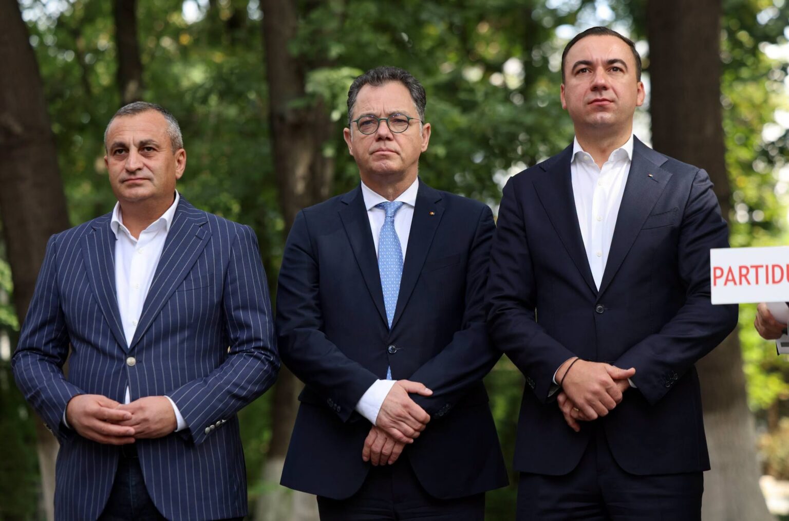 oprescu oprea ivan 841083411_1082498821293677_3410513739166288667_n - Jurnalul Olteniei – Știri din Craiova și Oltenia Three men in dark suits stand outdoors in a park, looking serious; one holds a sign with PARTI visible at the edge.