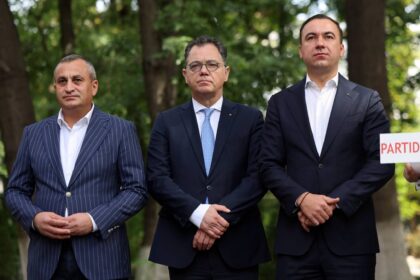 Three men in dark suits stand outdoors in a park, looking serious; one holds a sign with PARTI visible at the edge.