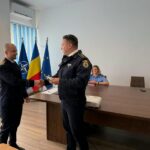 Two uniformed officers shake hands in a formal room with flags behind them and a woman seated at a desk nearby.