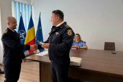 Two uniformed officers shake hands in a formal room with flags behind them and a woman seated at a desk nearby.