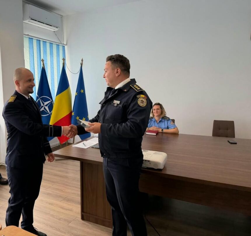Two uniformed officers shake hands in a formal room with flags behind them and a woman seated at a desk nearby.