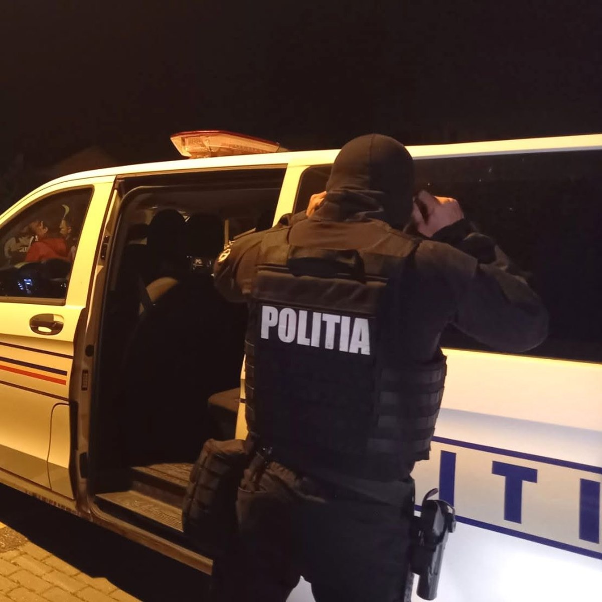 Officer in tactical gear standing by a police van at night, opening the side door to enter or check the vehicle.