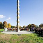 Tall vertical sculpture of stacked rounded blocks in a circular plaza, surrounded by people in a park on a sunny day.