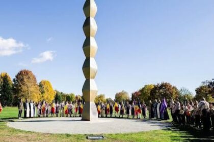 Tall vertical sculpture of stacked rounded blocks in a circular plaza, surrounded by people in a park on a sunny day.