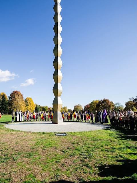 Tall vertical sculpture of stacked rounded blocks in a circular plaza, surrounded by people in a park on a sunny day.