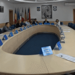 Large oval conference table with many people in a formal meeting, flags on the wall behind them