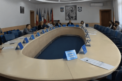 Large oval conference table with many people in a formal meeting, flags on the wall behind them