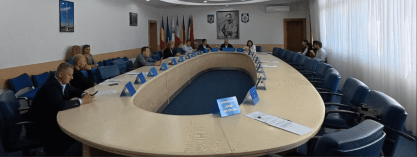 Large oval conference table with many people in a formal meeting, flags on the wall behind them
