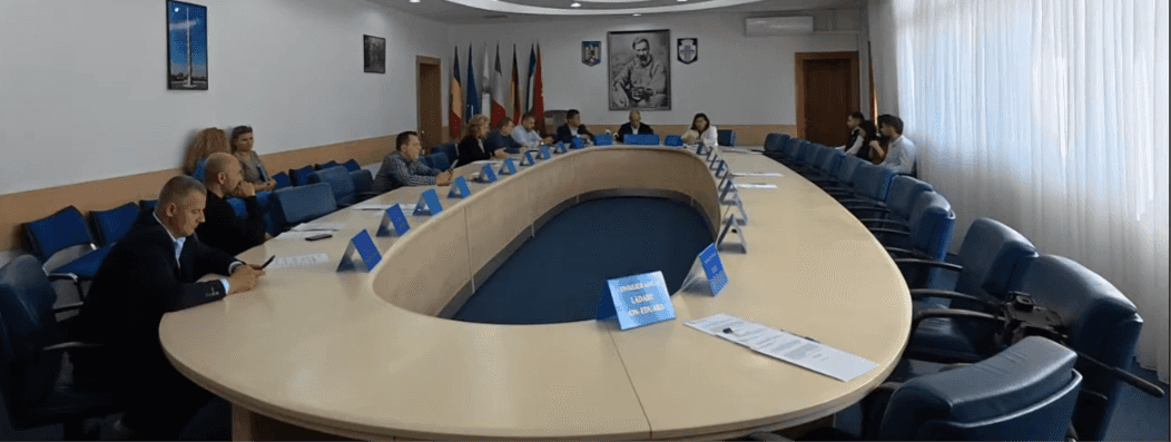 screenshot-2026-10-21-121438-8b84ab - Jurnalul Olteniei – Știri din Craiova și Oltenia Large oval conference table with many people in a formal meeting, flags on the wall behind them