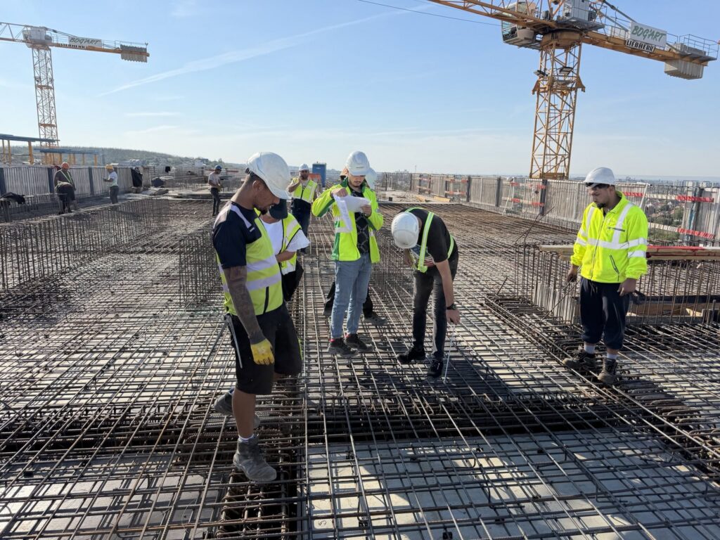 Construction workers wearing hard hats and high-visibility vests stand on a steel rebar grid at a high-rise construction site with cranes in the background.