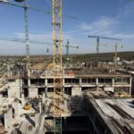 Construction site with tall green and yellow cranes over concrete building frames under a clear blue sky.