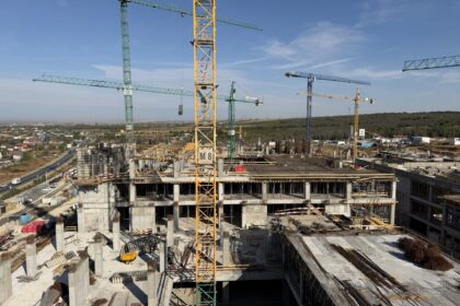 Construction site with tall green and yellow cranes over concrete building frames under a clear blue sky.