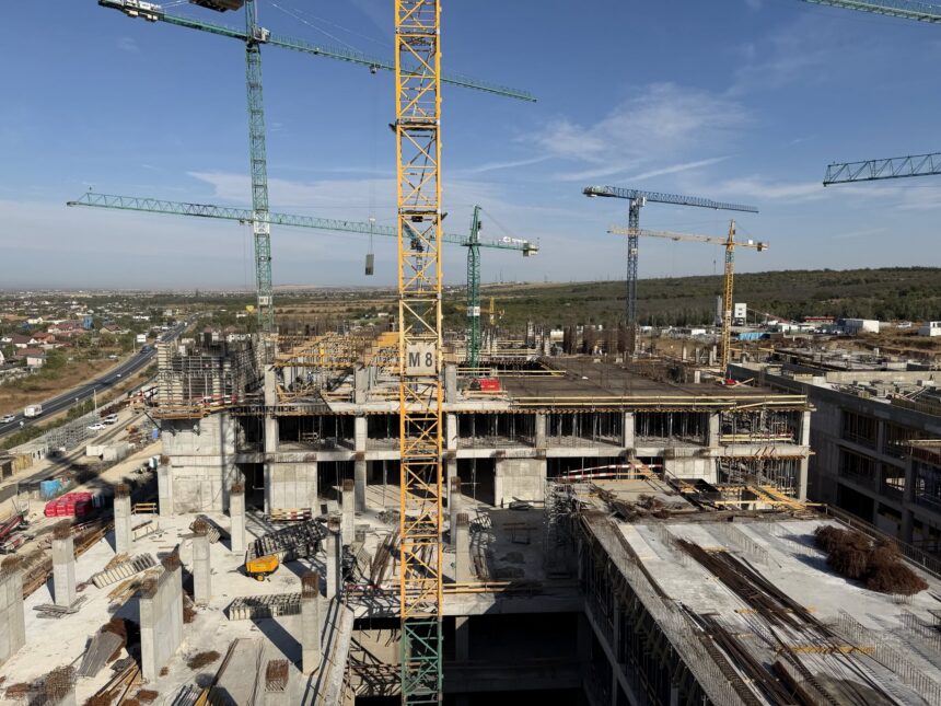 Construction site with tall green and yellow cranes over concrete building frames under a clear blue sky.