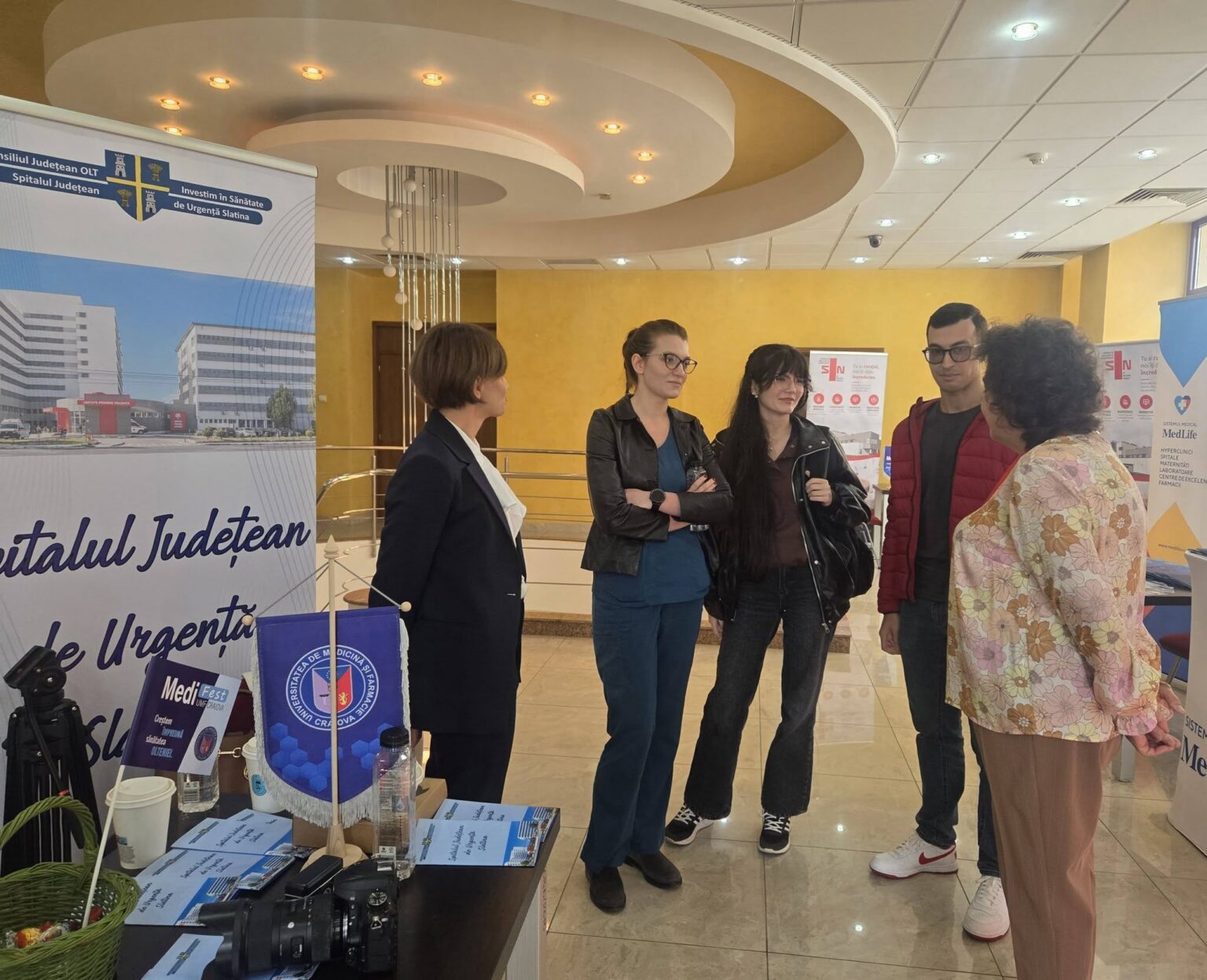 Group of people standing and talking at a medical information booth indoors. Posters and banners in the background show hospital branding.