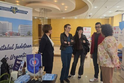 Group of people standing and talking at a medical information booth indoors. Posters and banners in the background show hospital branding.
